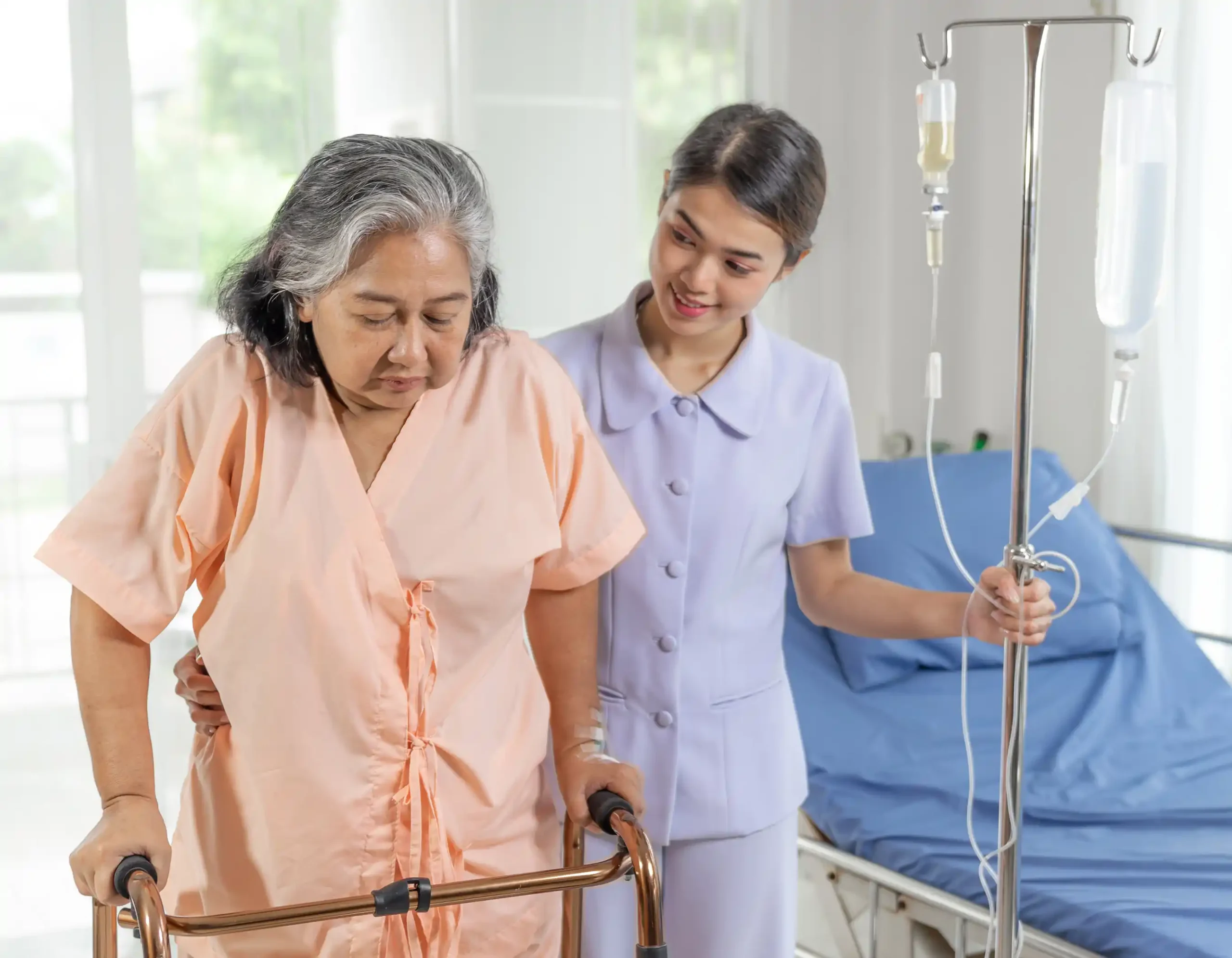 Older patient in peach hospital gown using walker assisted by healthcare worker in light purple uniform holding IV pole with two IV bags clear and yellow fluids, hospital bed with blue sheets in background representing patient mobility support and recovery care