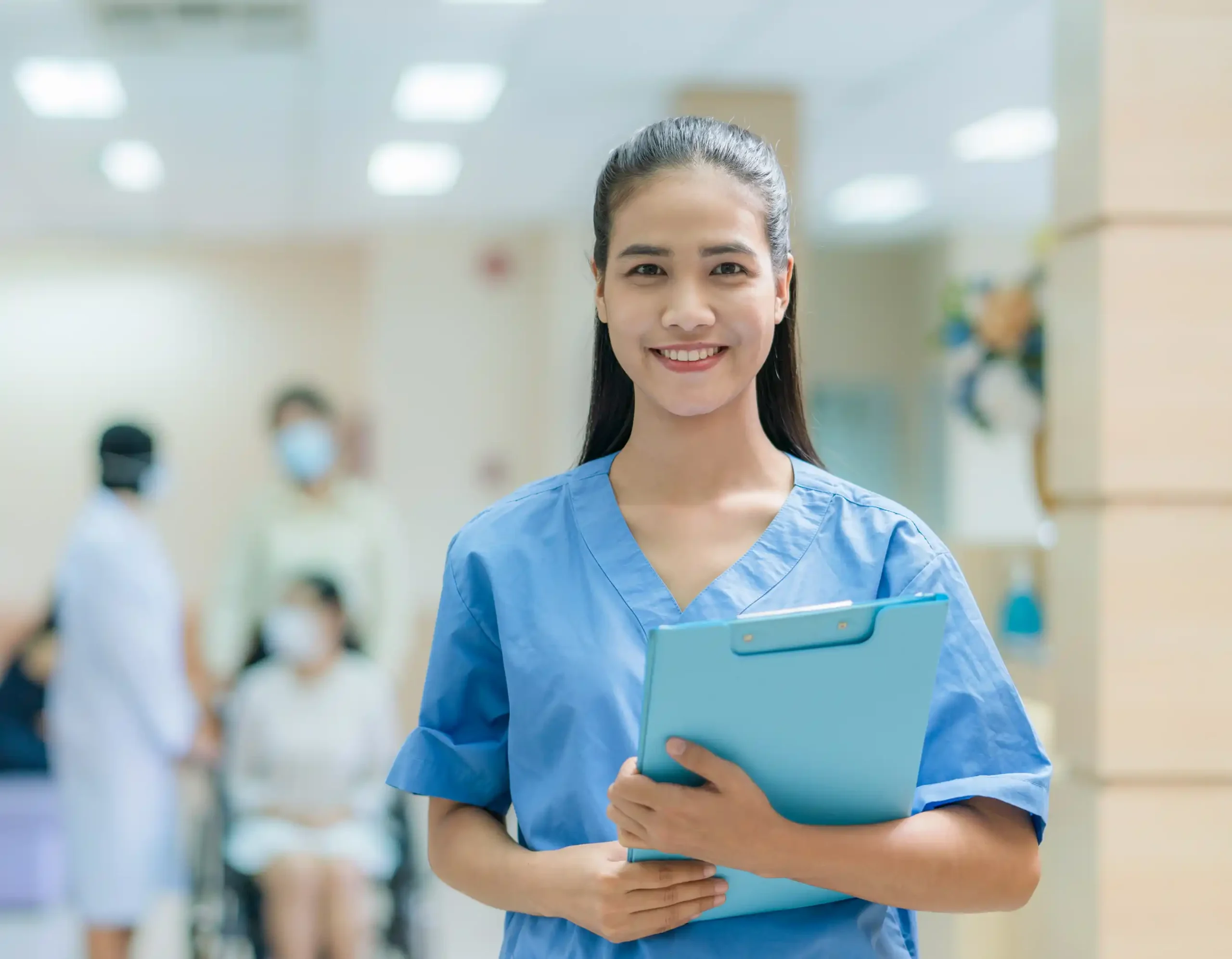 Healthcare professional in blue medical scrubs holding light blue clipboard standing in brightly lit hospital hallway with other staff in medical uniforms and face masks and patient seated in wheelchair in background