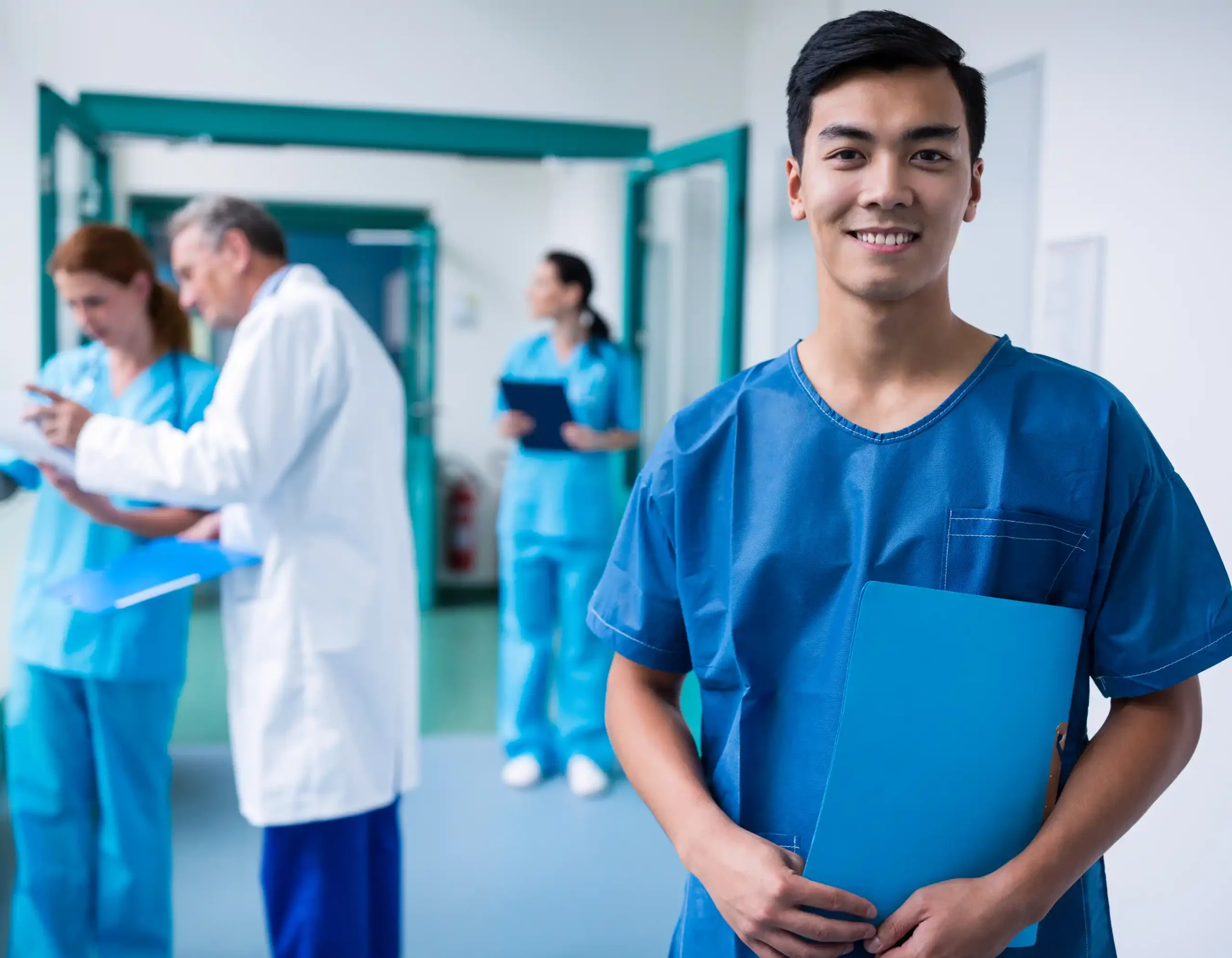 Group of healthcare professionals in hospital hallway including person in blue medical scrubs holding blue folder in foreground and three staff in medical attire two in blue scrubs and one in white lab coat reviewing documents together in clean brightly lit environment with teal doors
