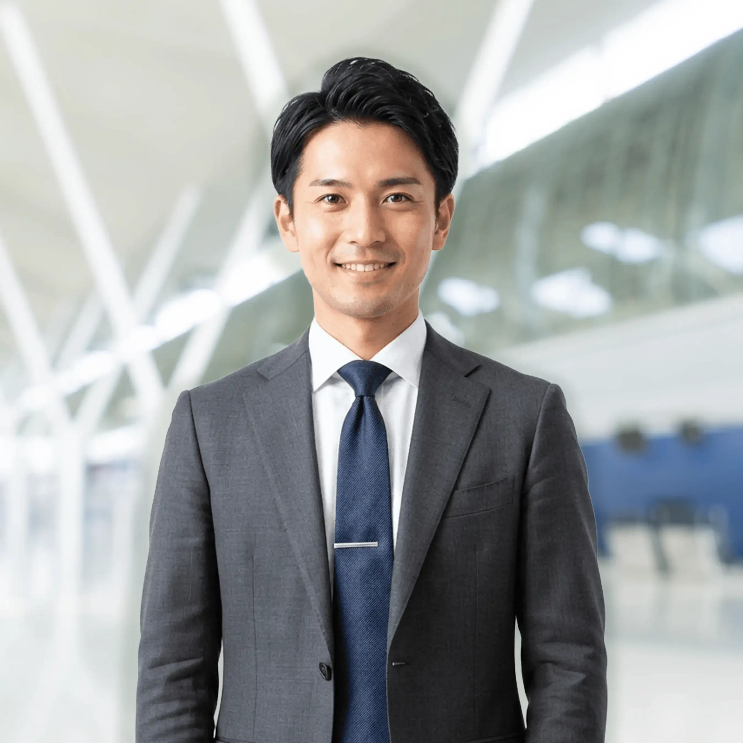 Person in dark gray suit white dress shirt and navy blue tie with silver tie clip standing indoors in modern bright space with large windows and white structural beams representing professional attire business environment and corporate setting