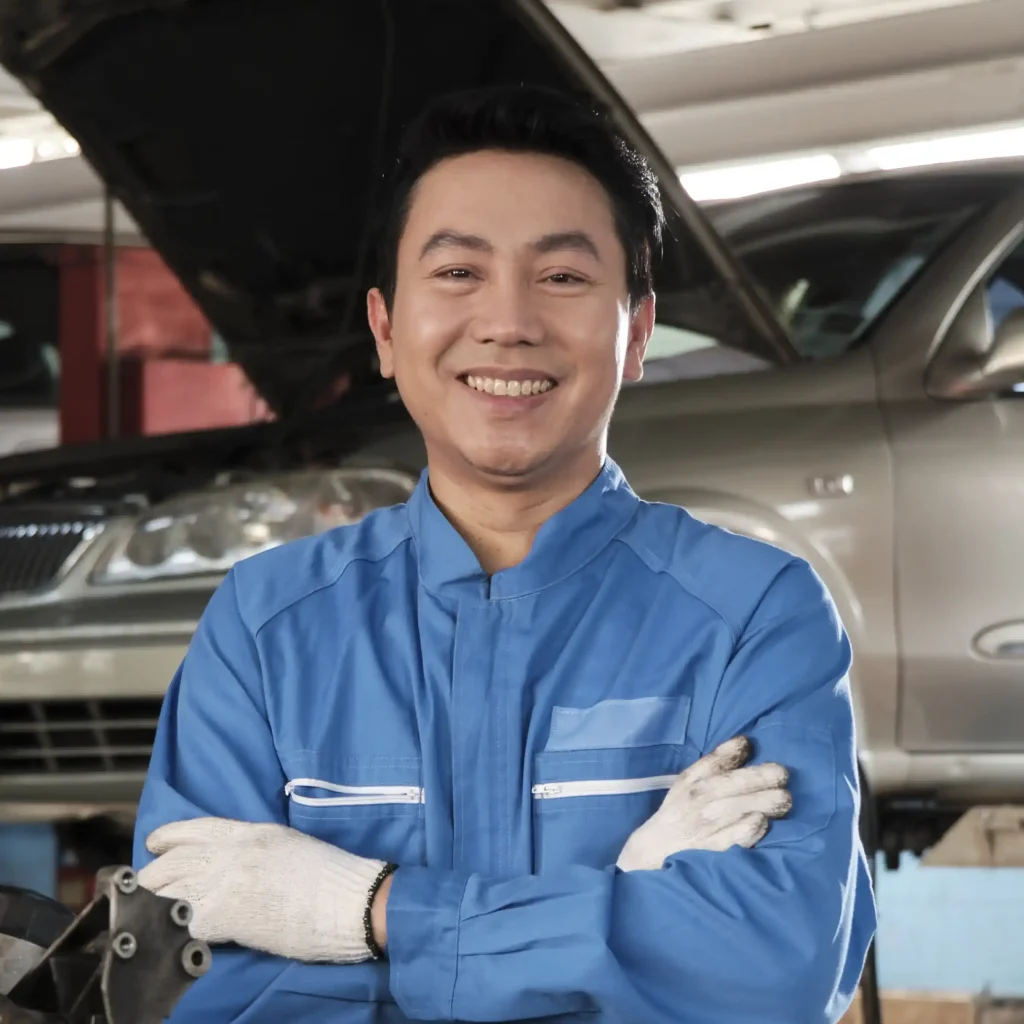 Mechanic in blue uniform and white gloves standing in front of car with hood open inside automotive repair shop showing front bumper headlights and raised hood representing vehicle inspection automotive maintenance and technical expertise