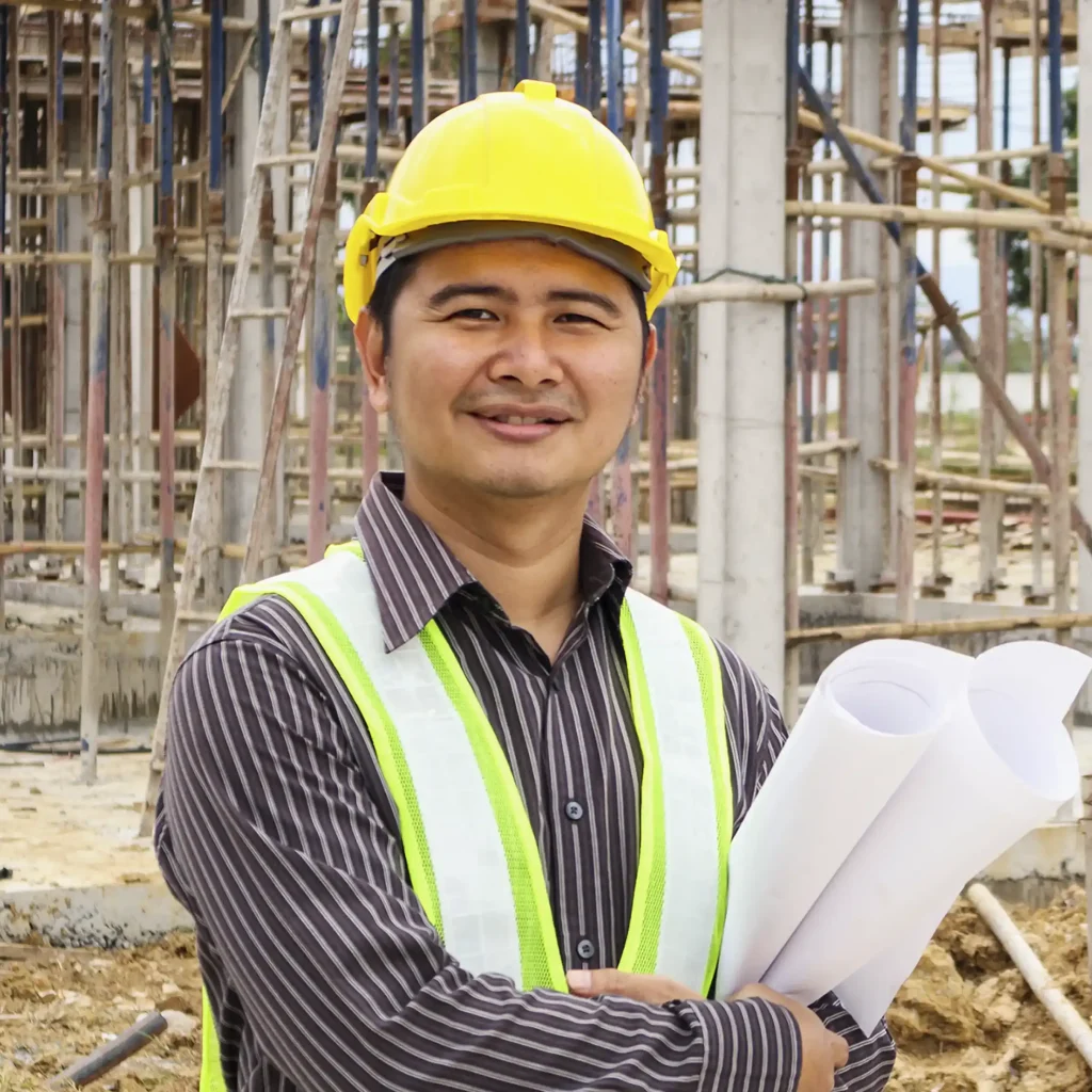 "Construction worker wearing yellow safety helmet and reflective vest holding rolled-up blueprints at active building site with scaffolding metal poles and partially constructed concrete columns representing engineering planning safety and project management