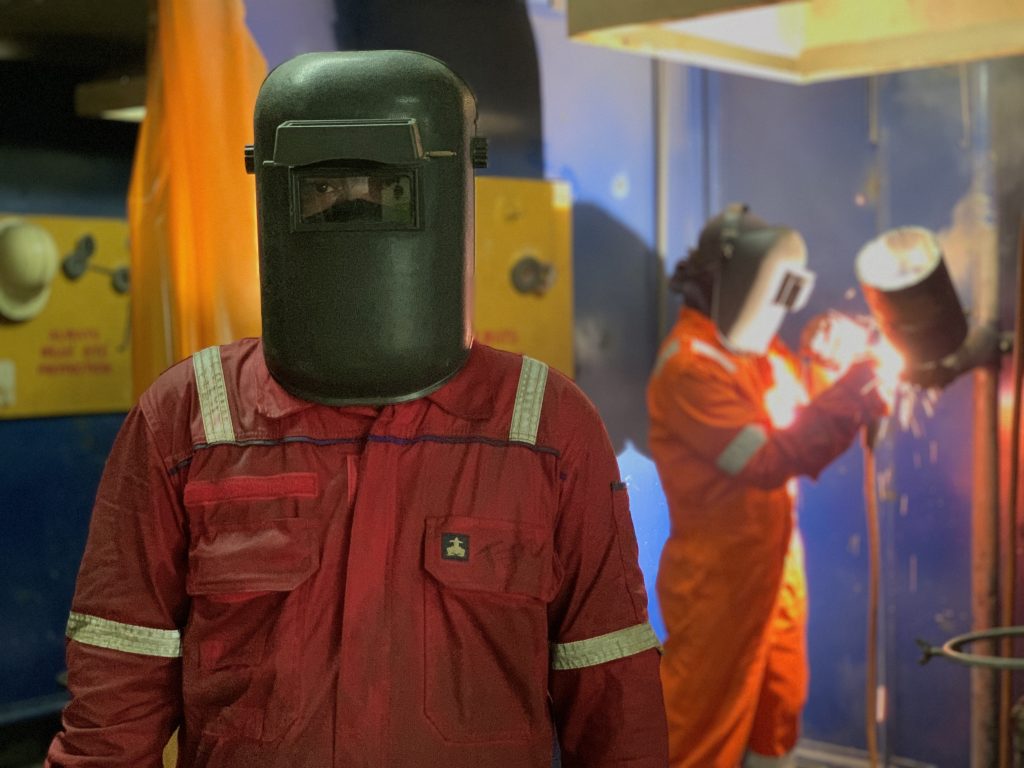 Two individuals in protective welding gear full-face helmets and flame-resistant coveralls one in red coverall facing camera and other in orange coverall actively welding metal piece with bright sparks in industrial workshop with blue and yellow walls and safety equipment representing welding operations and industrial safety practices