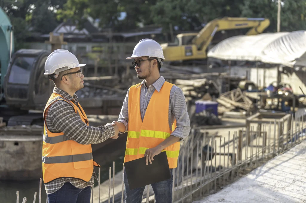 Two construction professionals wearing safety helmets and orange reflective vests shaking hands at active construction site with excavator steel rods and building materials in background one holding black folder representing professional agreement teamwork and project collaboration