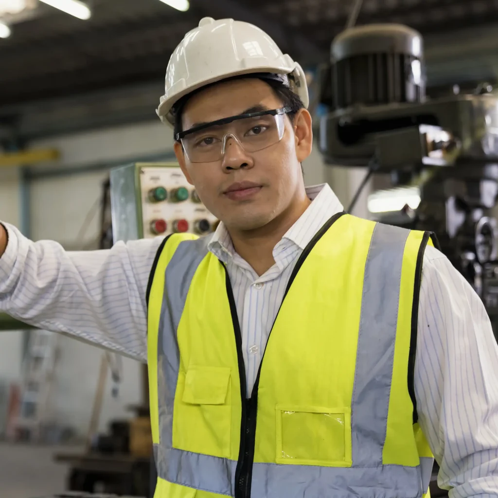 Person wearing white safety helmet clear protective glasses and high-visibility yellow vest with gray reflective strips standing in industrial environment with machinery and control panels in background representing occupational safety engineering construction and factory operations