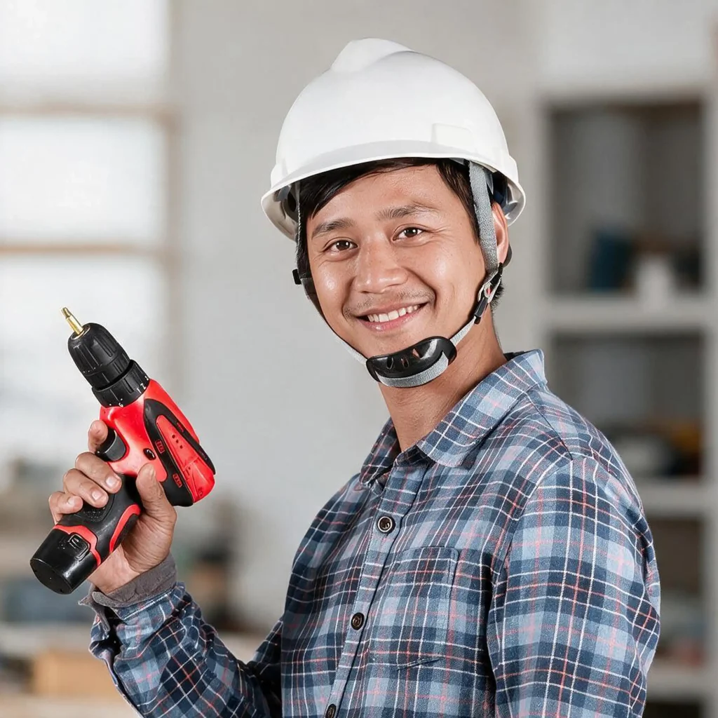 Person wearing white safety helmet and blue plaid shirt holding red and black cordless power drill with drill bit in indoor workshop with shelves and tools blurred in background representing construction DIY safety and use of power tools