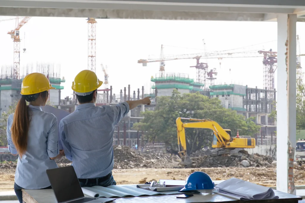 Two engineers in yellow safety helmets and light blue shirts standing at construction site pointing toward large building under construction with cranes scaffolding and concrete structures foreground showing table with blueprints laptop and blue helmet plus yellow excavator and dirt piles representing architectural planning engineering inspection and project progress